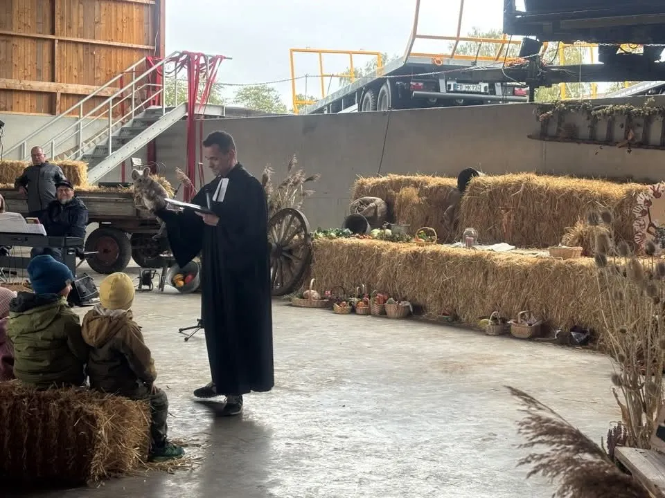 Der Pfarrer im Talar vor Strohballen mit Erntegaben, im Vordergrund Kinder auf Strohballen