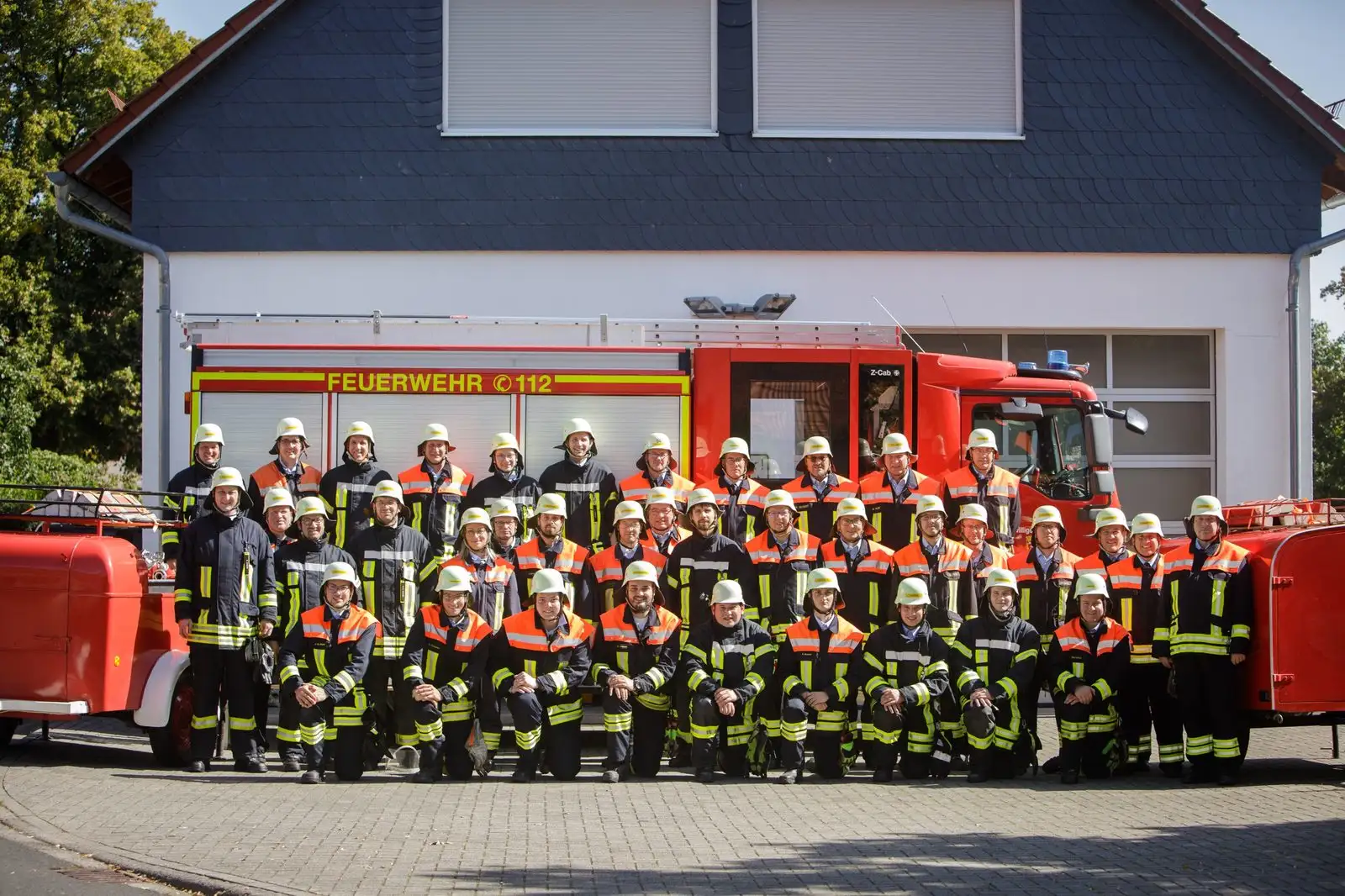 Die Einsatzabteilung in Schutzkleidung und Helm vor dem Feuerwehrhaus und einem roten Löschfahrzeug