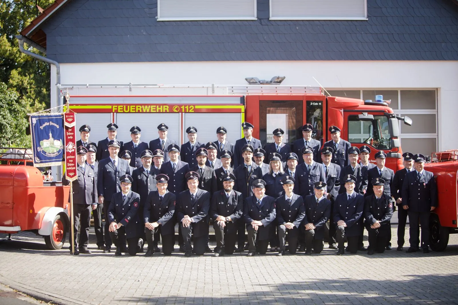 Die Einsatzabteilung in Uniform vor dem Feuerwehrhaus, links die Vereinsfahne