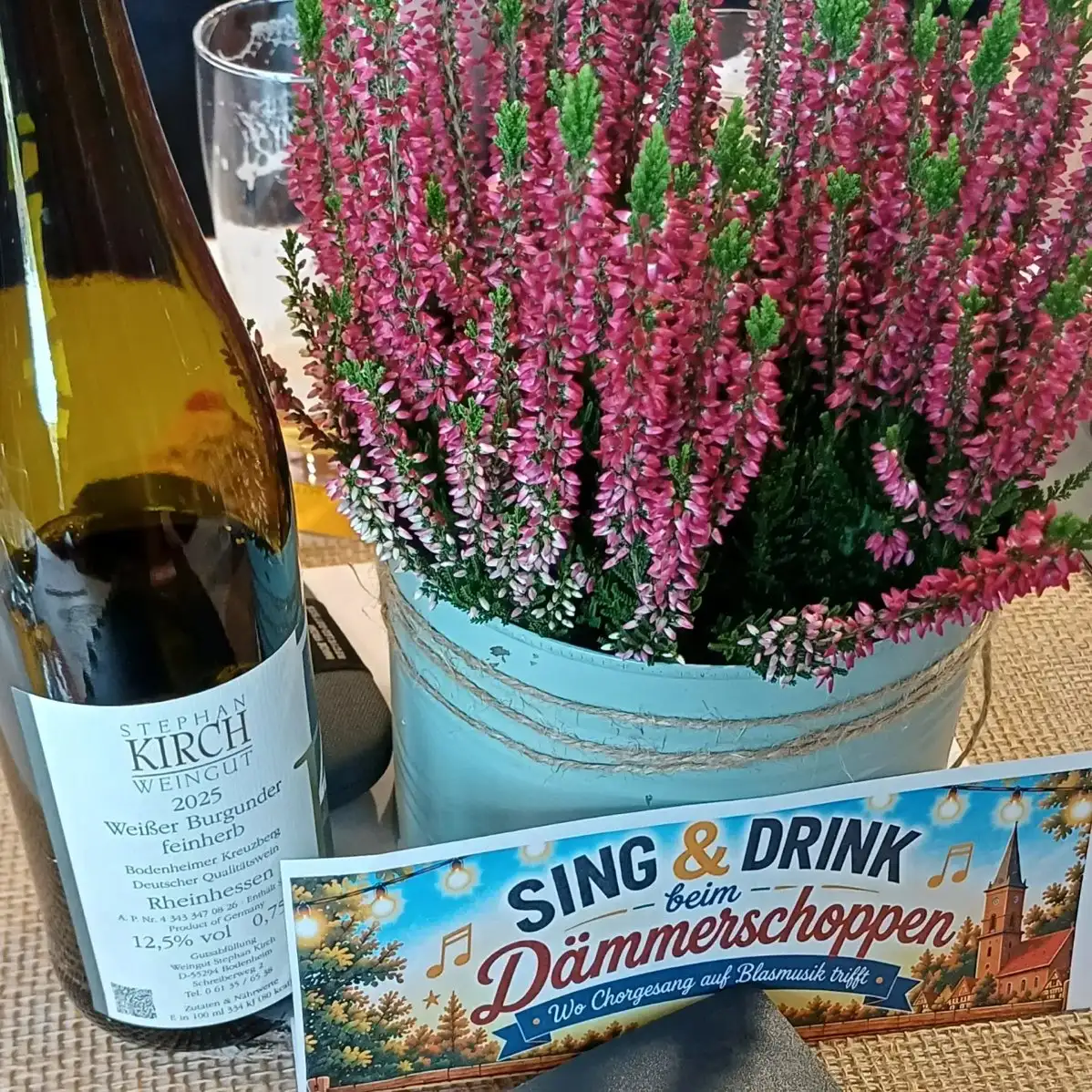 Weinflasche, Heidekraut im Topf und eine Karte „Sing & Drink beim Dämmerschoppen“ auf einem Tisch