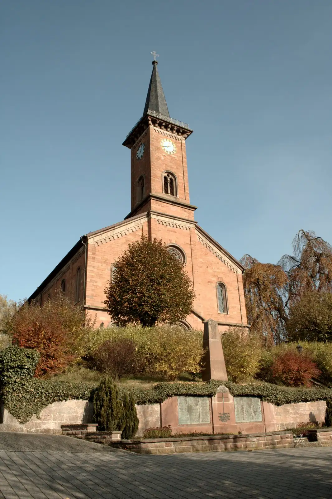 Die Kirche in Stockhausen: roter Sandsteinbau mit spitzem Turm und Uhr, davor Sträucher in Herbstfarben