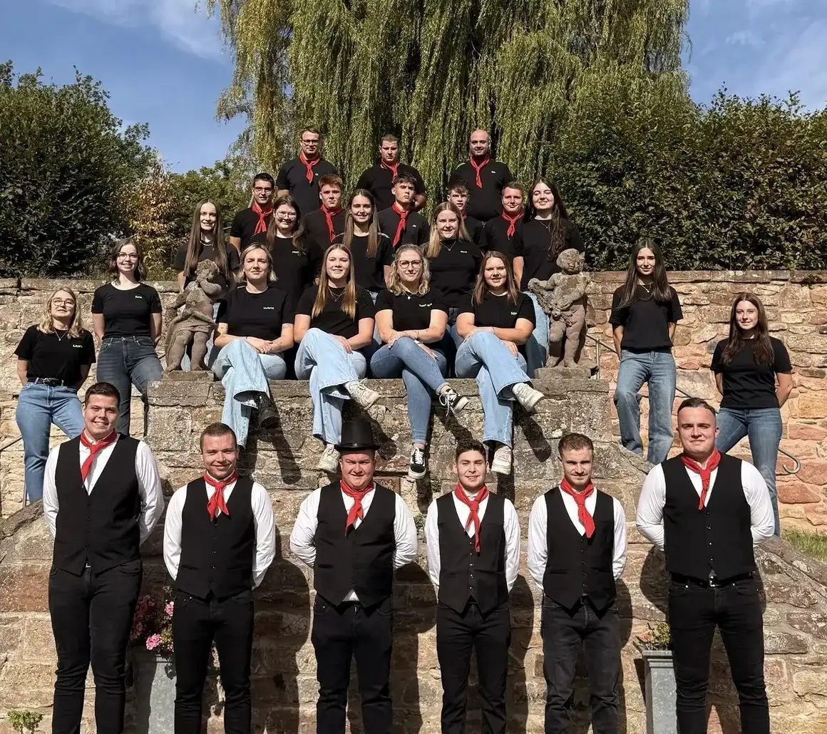 Gruppenfoto vor einer Sandsteinmauer: vorn sechs Kirmesburschen in schwarzen Westen mit roten Halstüchern, dahinter junge Leute in schwarzen T-Shirts