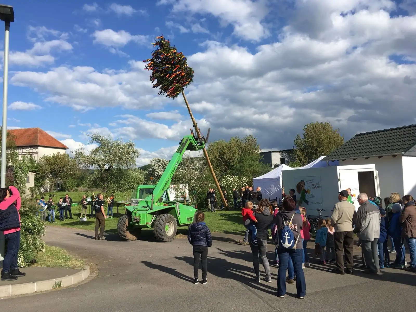 Ein geschmückter Maibaum wird mit einem grünen Teleskoplader aufgestellt, ringsum Zuschauer
