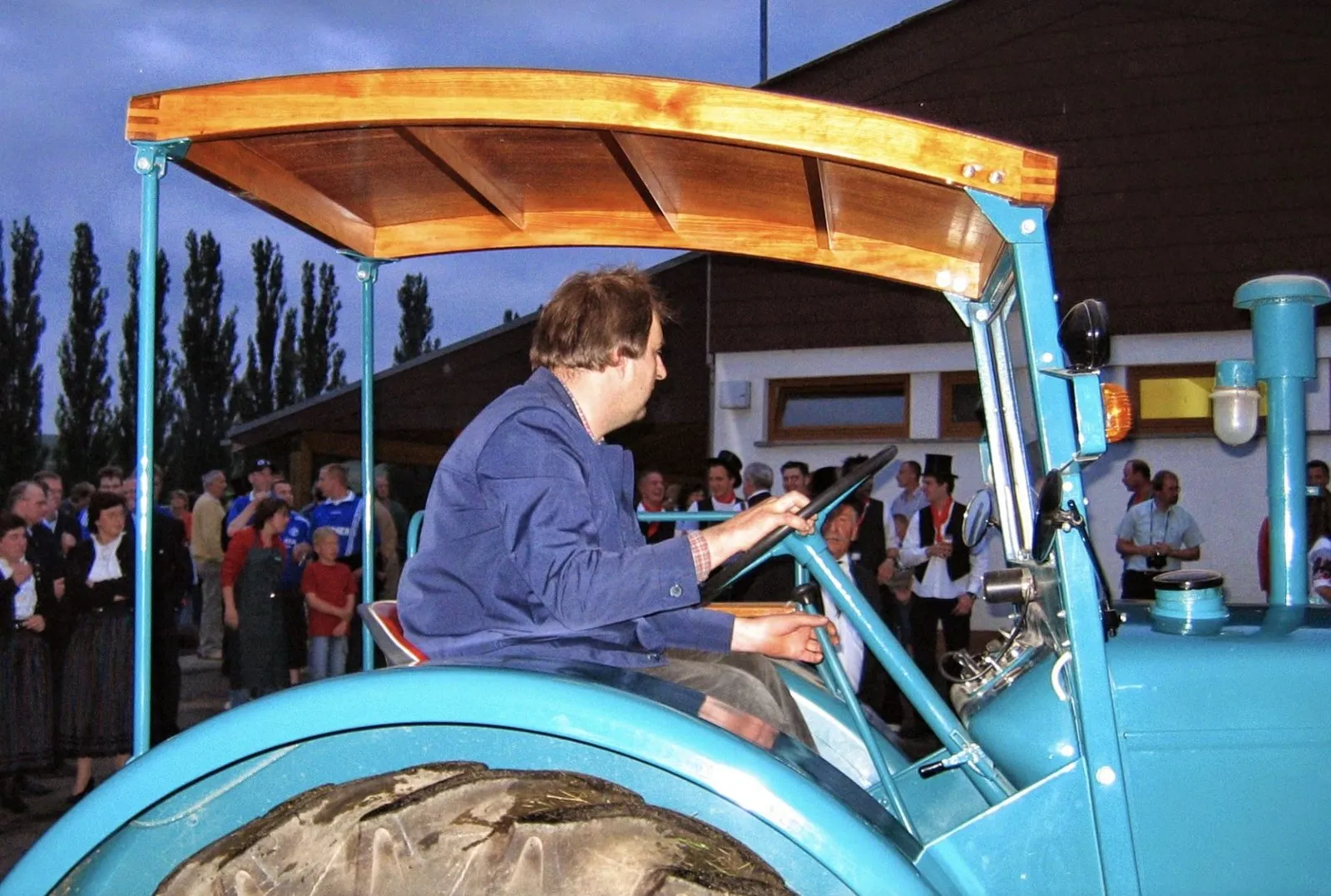 Fahrer am Steuer eines türkisfarbenen Hanomag mit Holzdach, im Hintergrund Zuschauer vor einer Halle