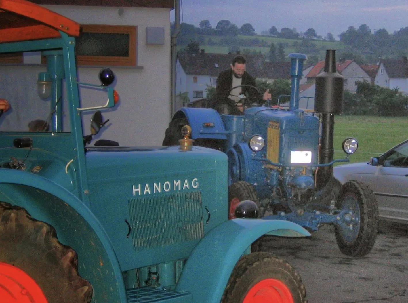Motorhaube eines türkisfarbenen Hanomag im Vordergrund, dahinter ein blauer Lanz Bulldog am Abend
