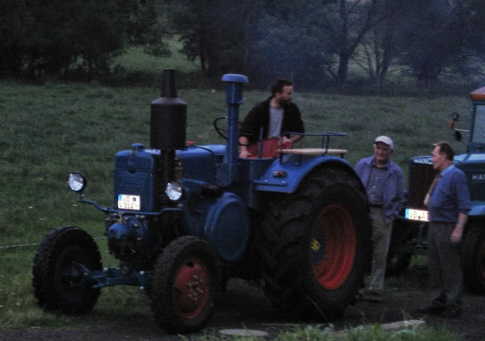Blauer Lanz Bulldog mit roten Felgen auf einer Wiese in der Abenddämmerung, daneben drei Männer im Gespräch