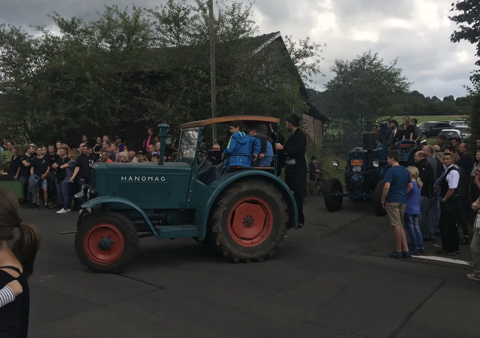 Grüner Hanomag mit Holzdach im Umzug durchs Dorf, dahinter ein blauer Lanz Bulldog, am Straßenrand viele Zuschauer