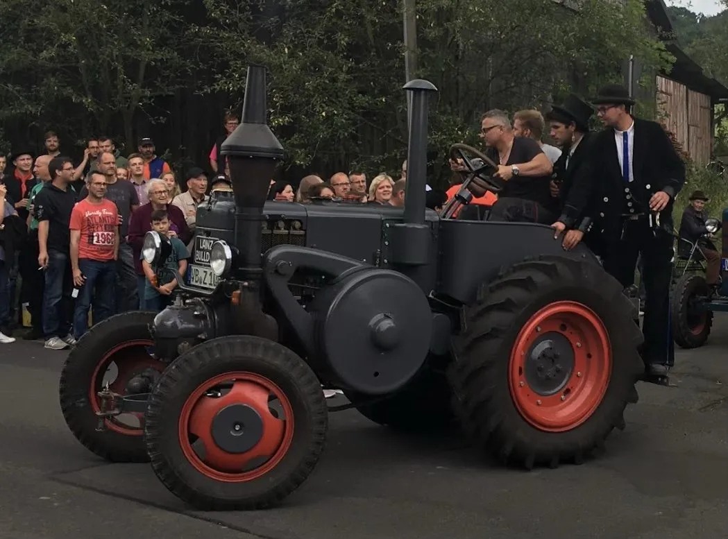 Grauer Lanz Bulldog mit roten Felgen und hohem Auspuff, Mitfahrende auf dem Kotflügel, im Hintergrund Zuschauer