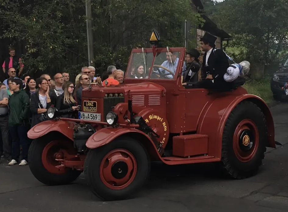 Roter Lanz Bulldog mit Windschutzscheibe, besetzt mit Mitfahrenden in Festkleidung, dahinter Zuschauer