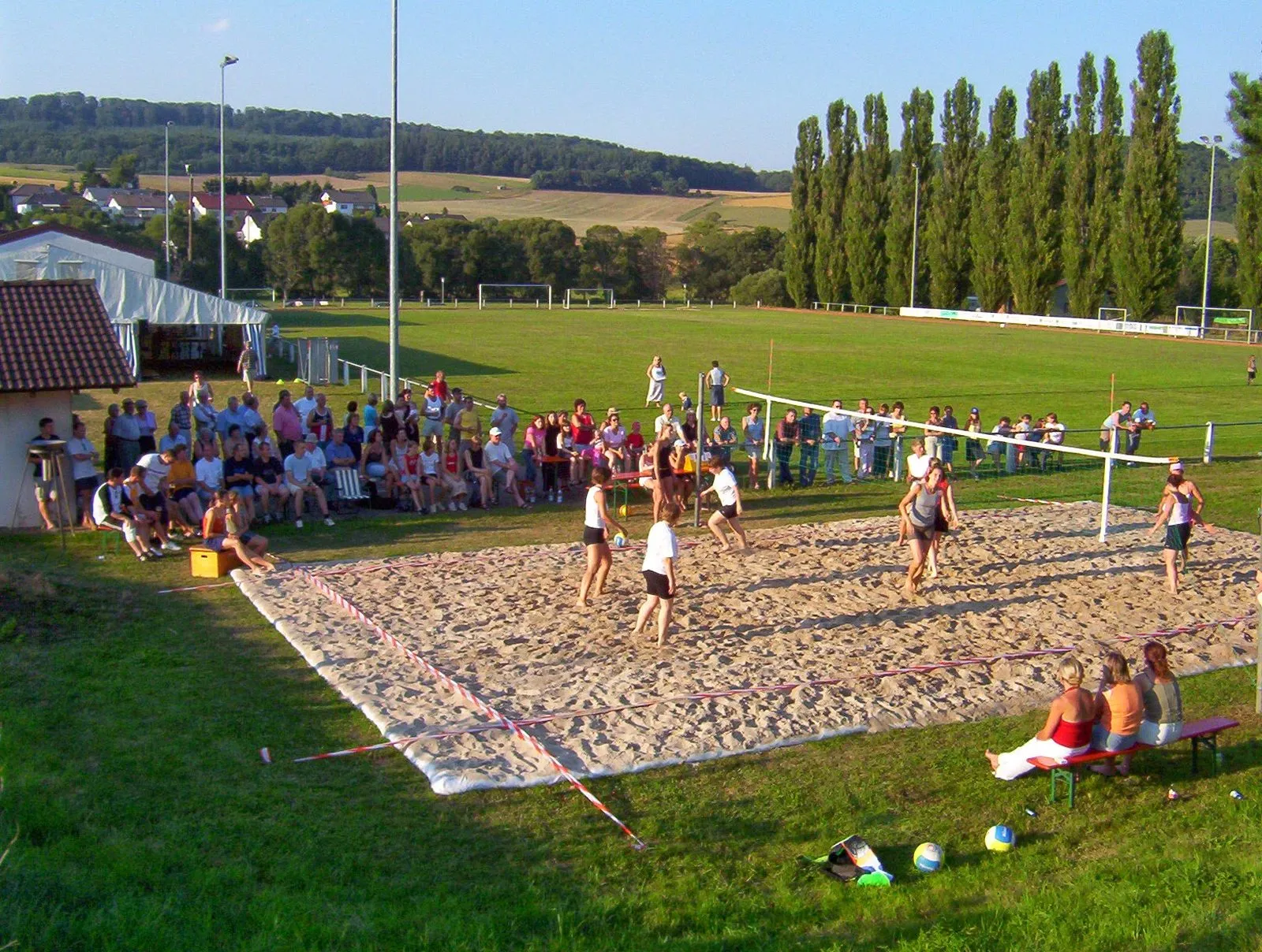 Beachvolleyball-Spiel im Sand am Sportplatz, ringsum viele Zuschauer, im Hintergrund das Fußballfeld und eine Pappelreihe