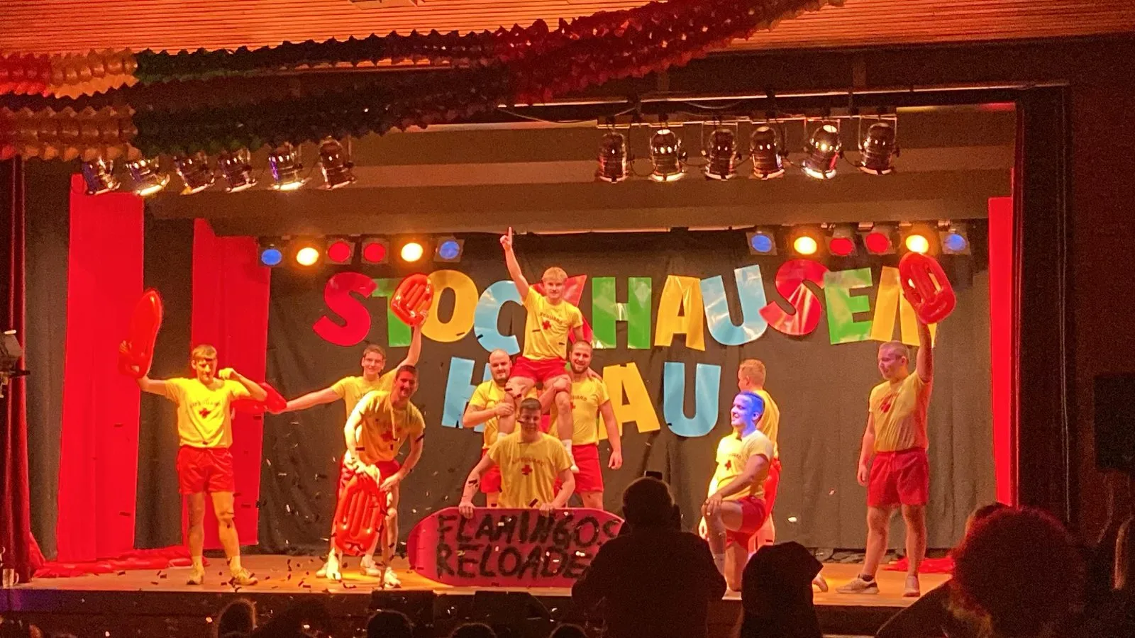 Faschingsauftritt auf der Bühne: Männergruppe als Rettungsschwimmer in gelben Shirts und roten Shorts vor dem bunten Schriftzug „Stockhausen Helau“, vorne ein Schild „Flamingos Reloaded“