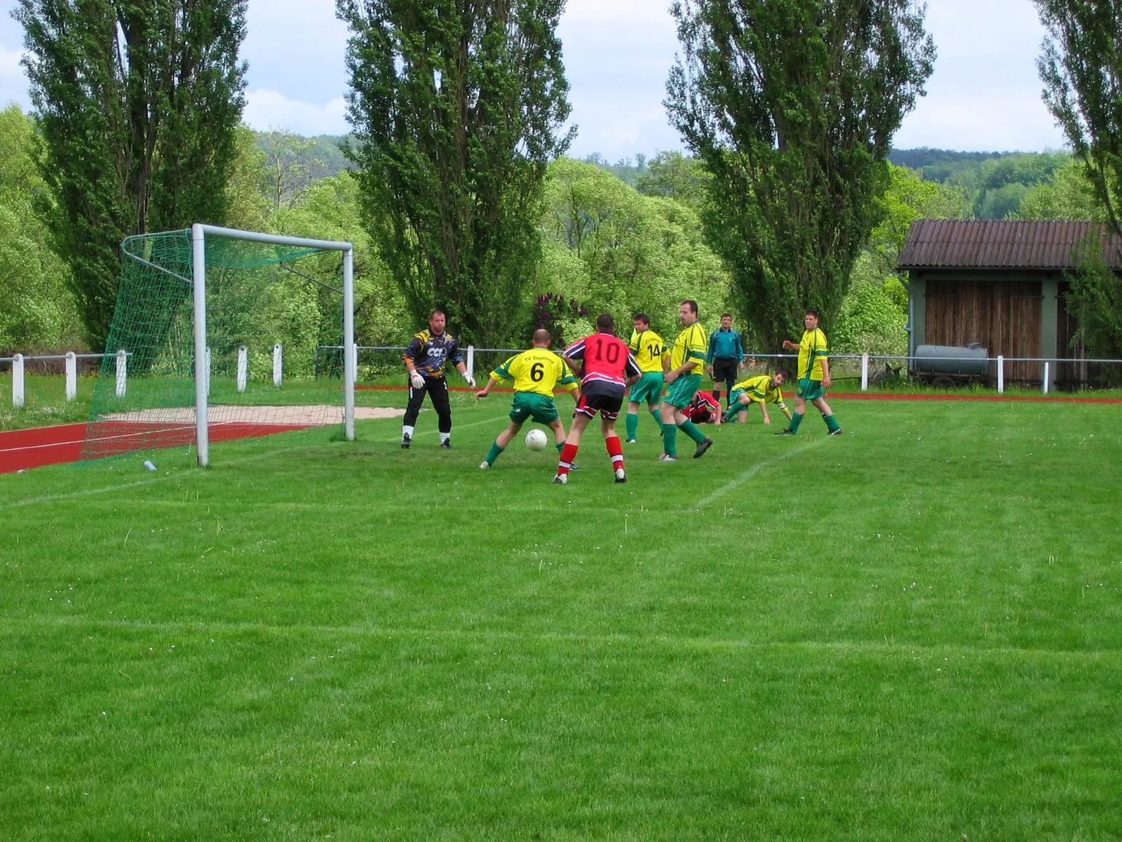Fußballspiel am Sportplatz: Ein Stürmer im roten Trikot vor dem Tor, umringt von Gegenspielern in Gelb, im Hintergrund Pappeln