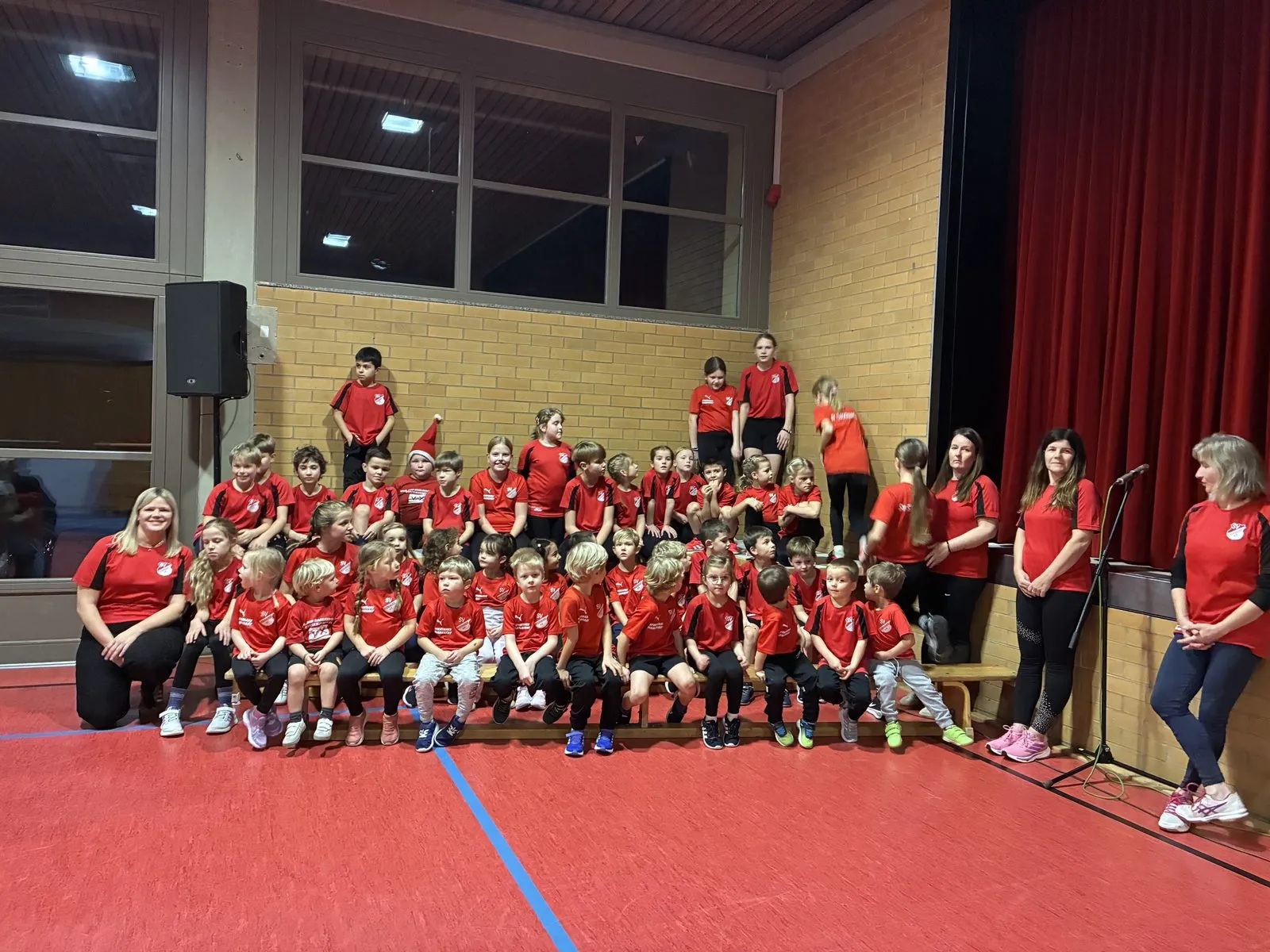 Die Turnkinder in roten SV-Trikots auf Bänken in der Halle, daneben das Trainerteam